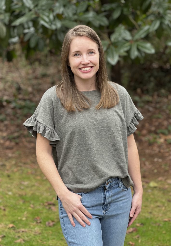 Brittany is standing outside, facing the camera. She has brown hair. She is wearing a green shirt and jeans.