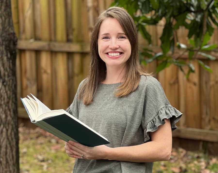 Brittany is holding a green and black book and smiling at the camera. She is standing outside and wearing a green shirt.