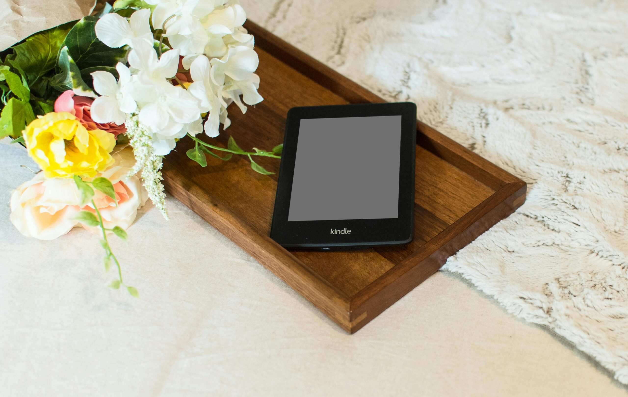 A kindle is sitting on a wooden tray with a bouquet of flowers next to it.